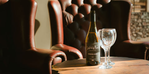 A bottle of Hammered Heron wine and two wine glasses resting on a wooden table in front of buttoned leather armchairs.