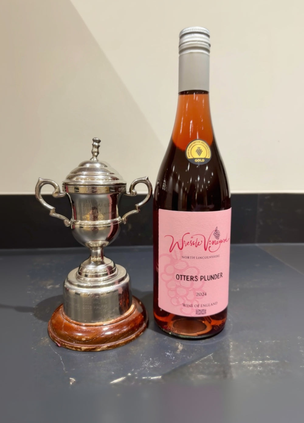 Bottle of Wressle Vineyard Otters Plunder 2024 rose wine next to a silver trophy on a marble worktop.