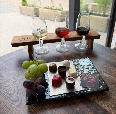 Three wine glasses with different colored wines on a wooden stand, accompanied by fruits and a chocolate assortment on a tray.