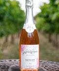 A bottle of Wressle Vineyard Dear Roe Deer 2024 sparkling rose wine stands on a metal table outdoors with a blurred vineyard background. The bottle has a white label with pink floral accents.