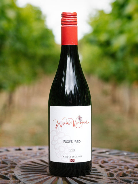 A bottle of Wressle Vineyard Foxed Red 2025 wine stands on a decorative metal table with a blurred outdoor vineyard background.