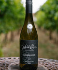 A bottle of Wressle Vineyard Hammered Heron 2024 white wine stands on a decorative metal table in a vineyard with blurred green grapevines in the background.