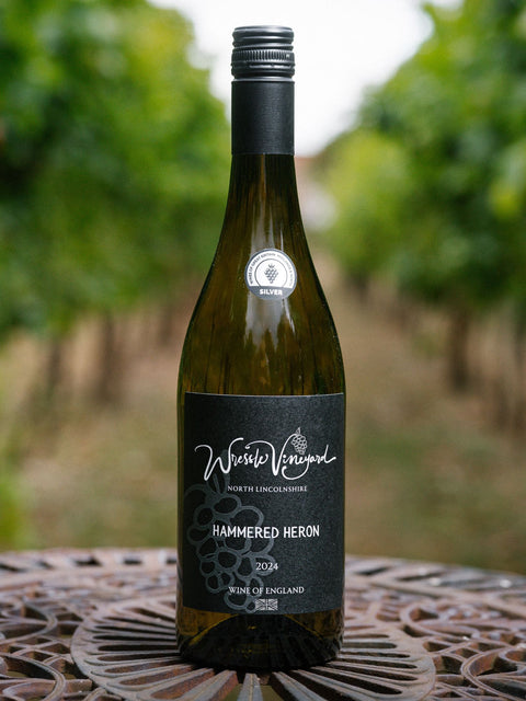 A bottle of Wressle Vineyard Hammered Heron 2024 white wine stands on a decorative metal table in a vineyard with blurred green grapevines in the background.