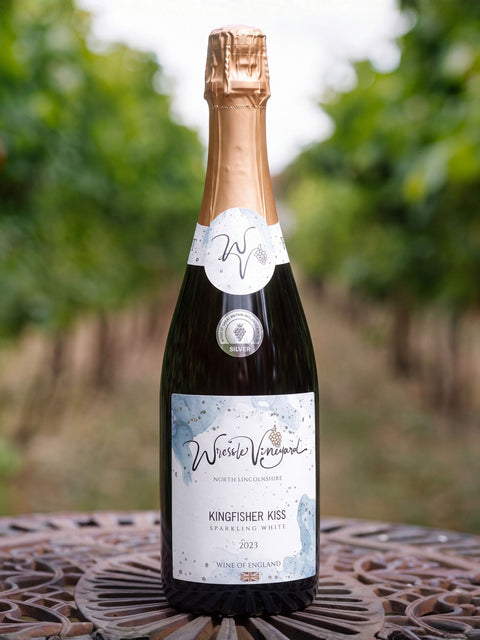 A bottle of Wressle Vineyard Kingfisher Kiss 2023 Sparkling White wine stands on a metal patio table with a blurred vineyard background.