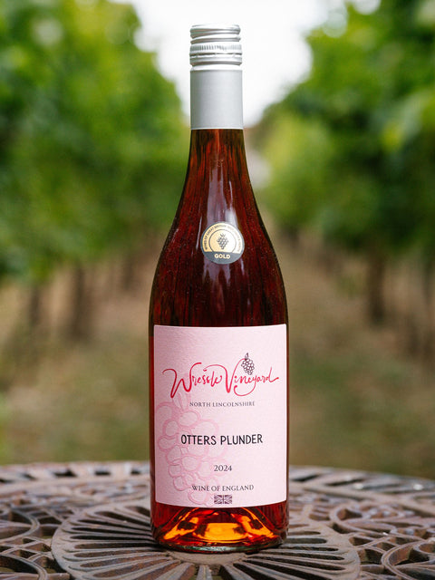 A bottle of Wressle Vineyard Otters Plunder 2024 rose wine stands on a metal patio table with a blurred vineyard background.