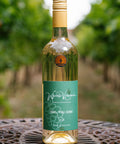 A bottle of Wressle Vineyard Owl You Doin' 2024 white wine stands on a decorative metal table in a vineyard with blurred green grapevines in the background.