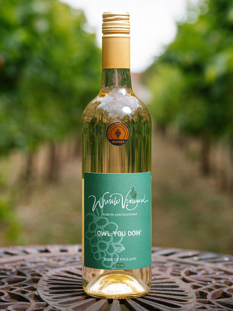 A bottle of Wressle Vineyard Owl You Doin' 2024 white wine stands on a decorative metal table in a vineyard with blurred green grapevines in the background.