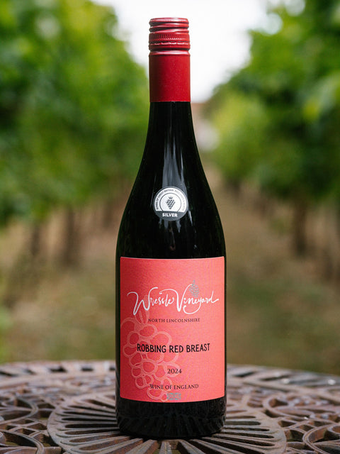 A bottle of Wressle Vineyard Robbing Red Breast 2024 red wine stands on a metal patio table with a blurred vineyard background.