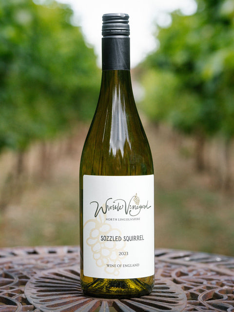 A bottle of Wressle Vineyard Sozzled Squirrel 2023 wine stands on a decorative metal table with a blurred vineyard background.