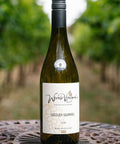 A bottle of Wressle Vineyard Sozzled Squirrel  2024 white wine stands on a decorative metal table in a vineyard with blurred green grapevines in the background.